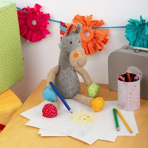 Sigikid plišasti konjiček sedi na mizi ob otroški risbi sonca in barvnih svinčnikih.